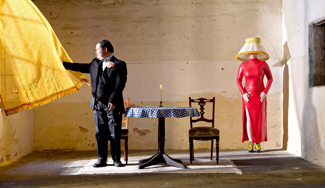 A man wearing a tuxedo looks out a window with a yellow curtain. A table, chair and mannequin with a lampshade in the background.