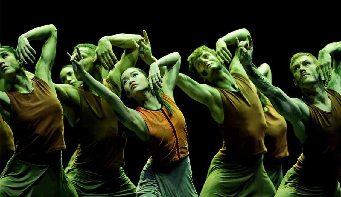 A group of dancers pose with one arm outstretched. They are all wearing orange tank tops and are bathed in a greenish light.