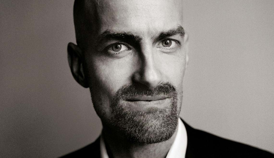 A black and white photo of Anthony Romaniuk, a bald man with a beard. He is wearing a white shirt and dark jacket and looks at the camera with a half-smile.