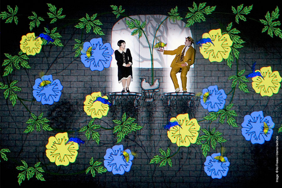 A lady and a man stand on a balcony each while blue and yellow flowers fall around them