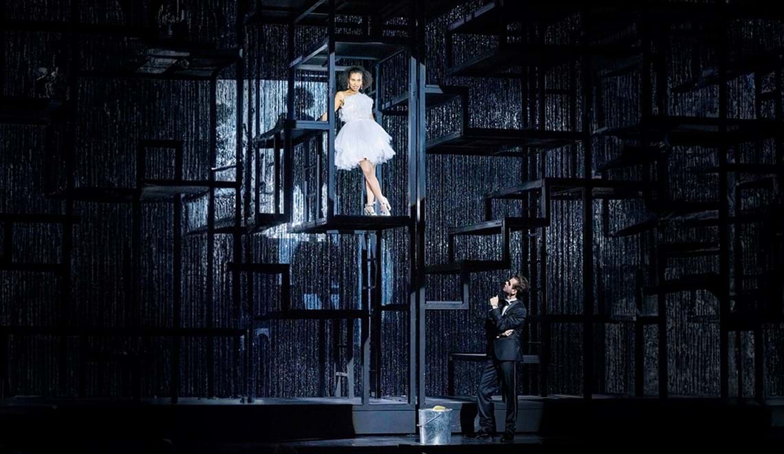 A woman in a white, tutu-like dress stands on a black structure in front of a silver-spangled backdrop while a man in a suit stands on the stage looking up at her.