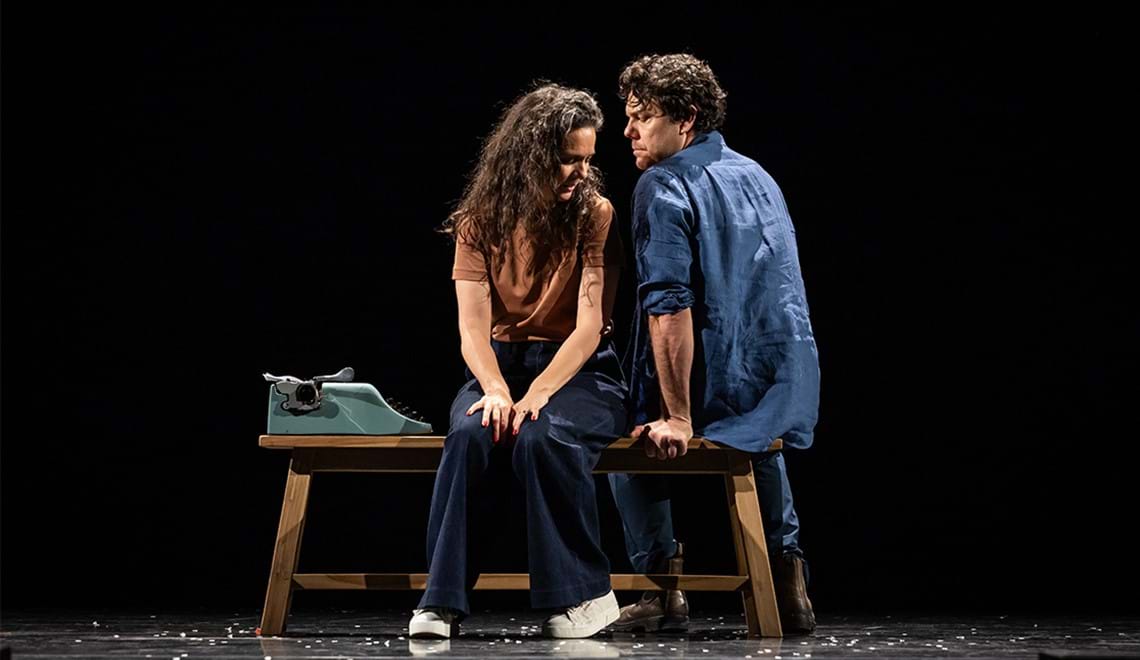Two people sit on a bench next to a type writer. One is wearing a blue shirt and has short brown hair and the other has an orange shirt with long curly brown hair. They are sitting facing opposite directions but face each other with serious expressions.