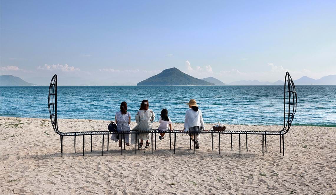 Four people sit on a forged steel bench facing away from the camera. The bench is on the beach and the background is a view of the ocean with land masses across the water.