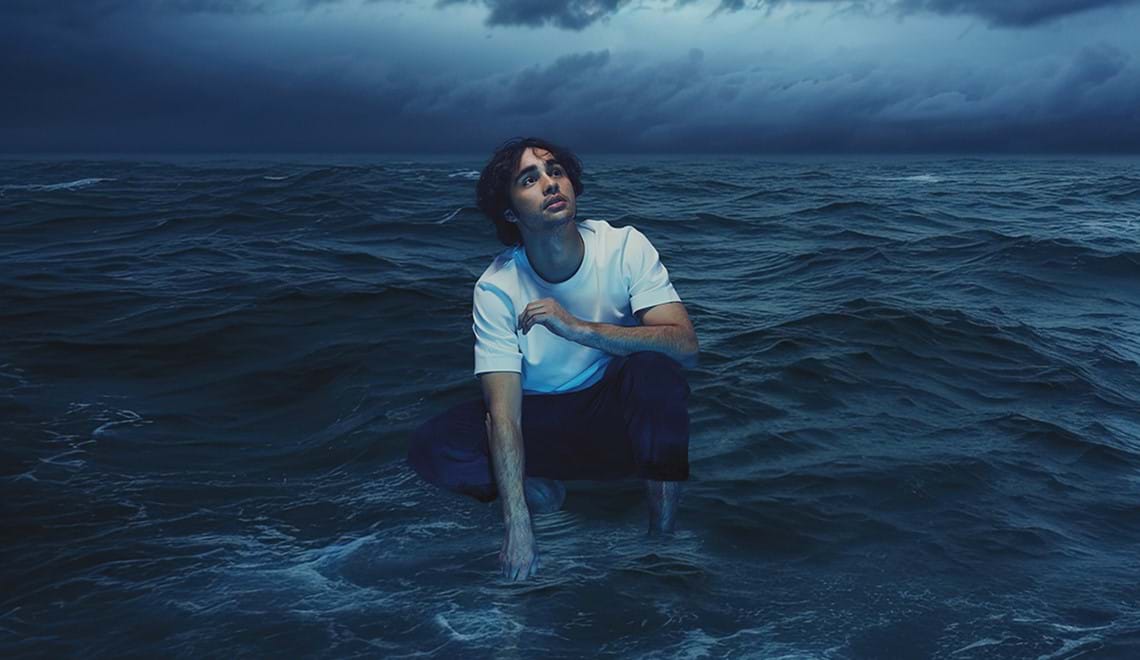 A young man with dark wavy hair and wearing a white t-shirt is pictured crouching down superimposed on a background of an ocean and stormy sky in shades of blue.