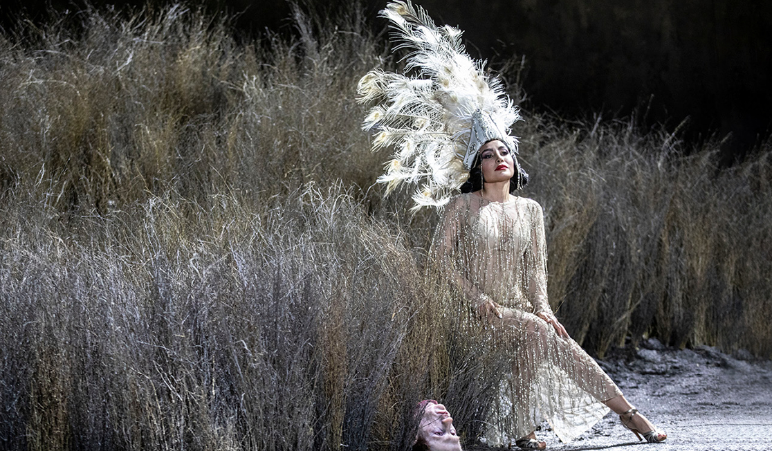 A beautiful woman, wearing a feathered headpiece and a 1920's style dress, sits in a field with tall, dry grass. The head of a dead man lying on the ground next to her.