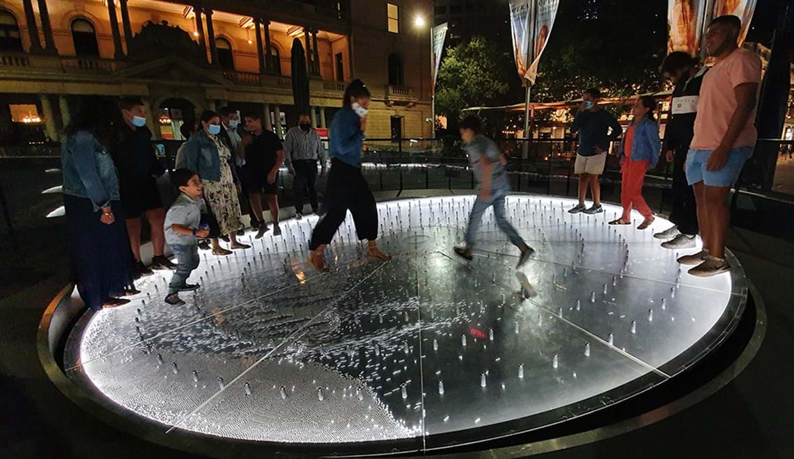 People stand around the edges of a silver disc that's tilted on an angle. Three people are walking through the centre and are the cause of the tilt.