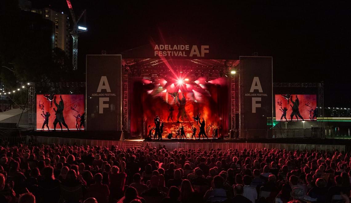 A group of people perform acrobatic moves on a large stage with Adelaide Festival branding in front of a large crowd.