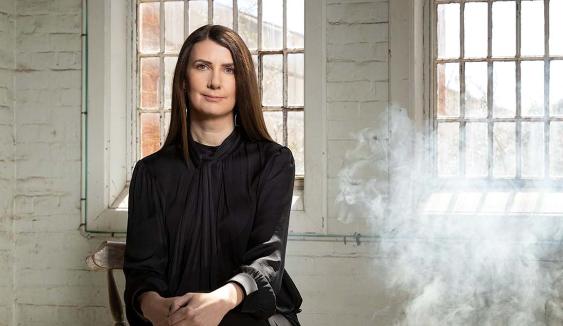 Anna Goldsworthy, a white woman with straight brown hair, sits in front of a white brick wall and window. She wears black and her hands are resting on her knee.