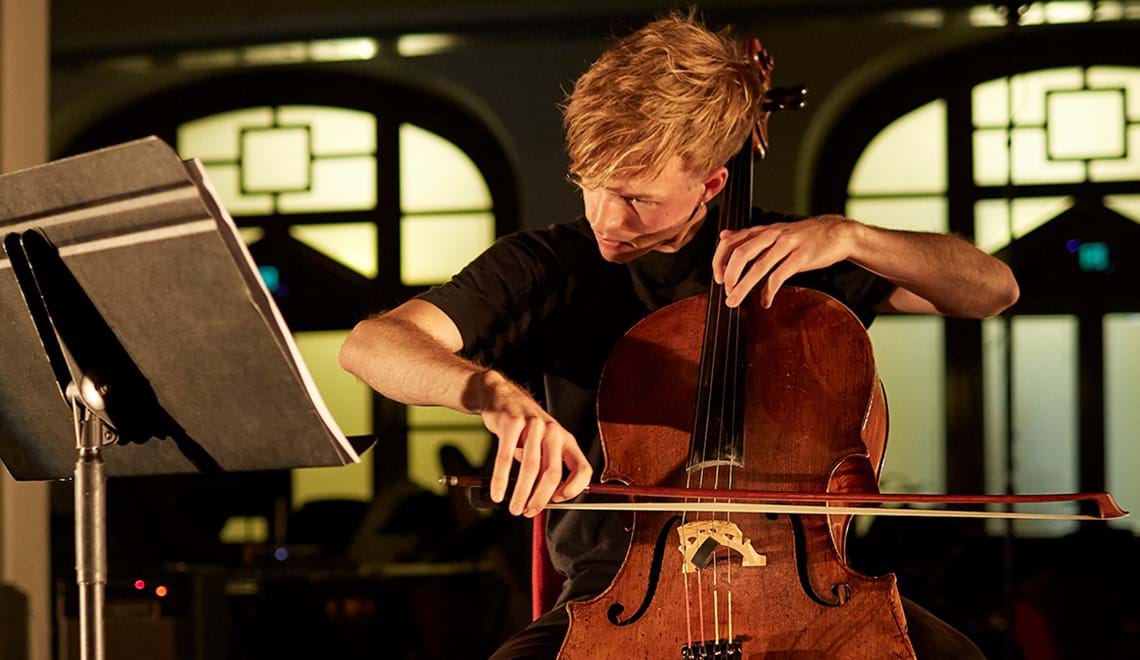 James Morley, a young man with side-parted blond hair and wearing a black t-shirt, is seated and playing the cello.