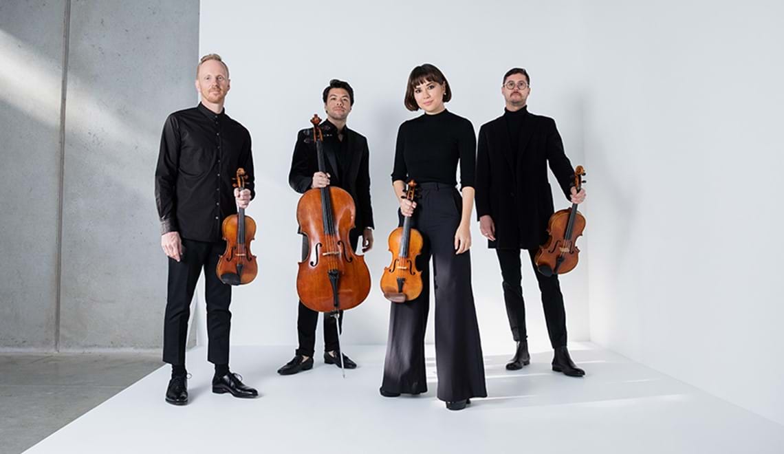 Four musicians dressed in black stand on a white floor with a white wall behind them. They are all holding string instruments.