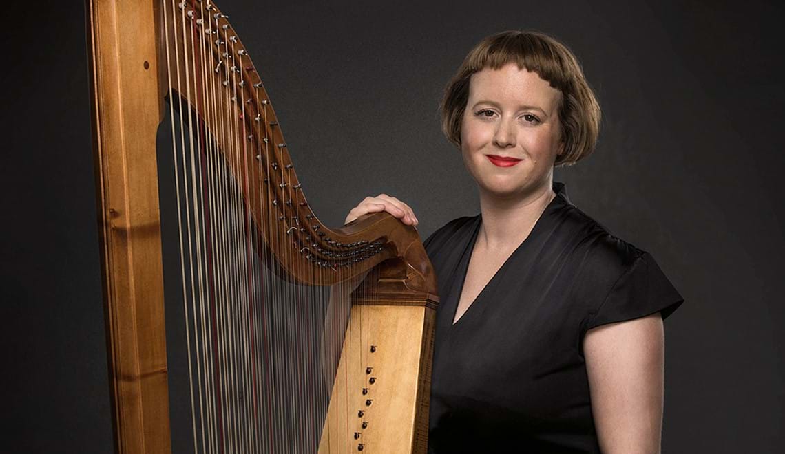 Hannah Lane, a woman with short brown hair wearing red lipstick and a black dress, stands with a baroque harp in front of a black background.
