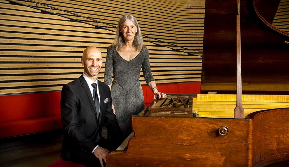 Erin Helyard is wearing a black suit and sitting next to an 1853 Érard piano. Stephanie McCallum stands behind him, wearing a silver dress.
