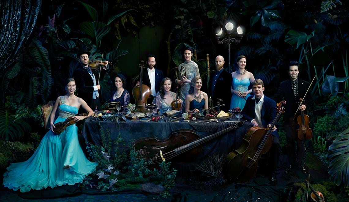 Members of the Australian Haydn Ensemble are seated around a dinner table draped in fabric and laden with fruit in a dark, leafy setting.  They are wearing formalwear and are holding their instruments.
