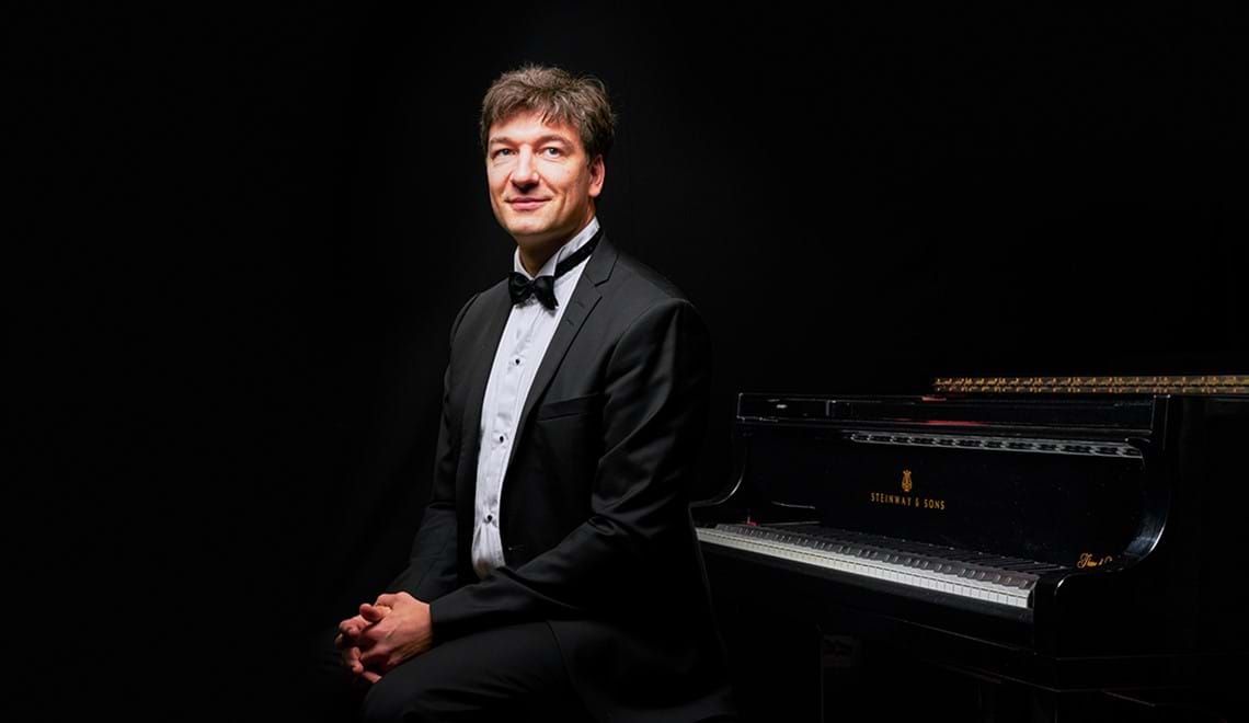 Konstantin Shamray wears a tuxedo and sits next to a piano with his hands clasped in his lap.