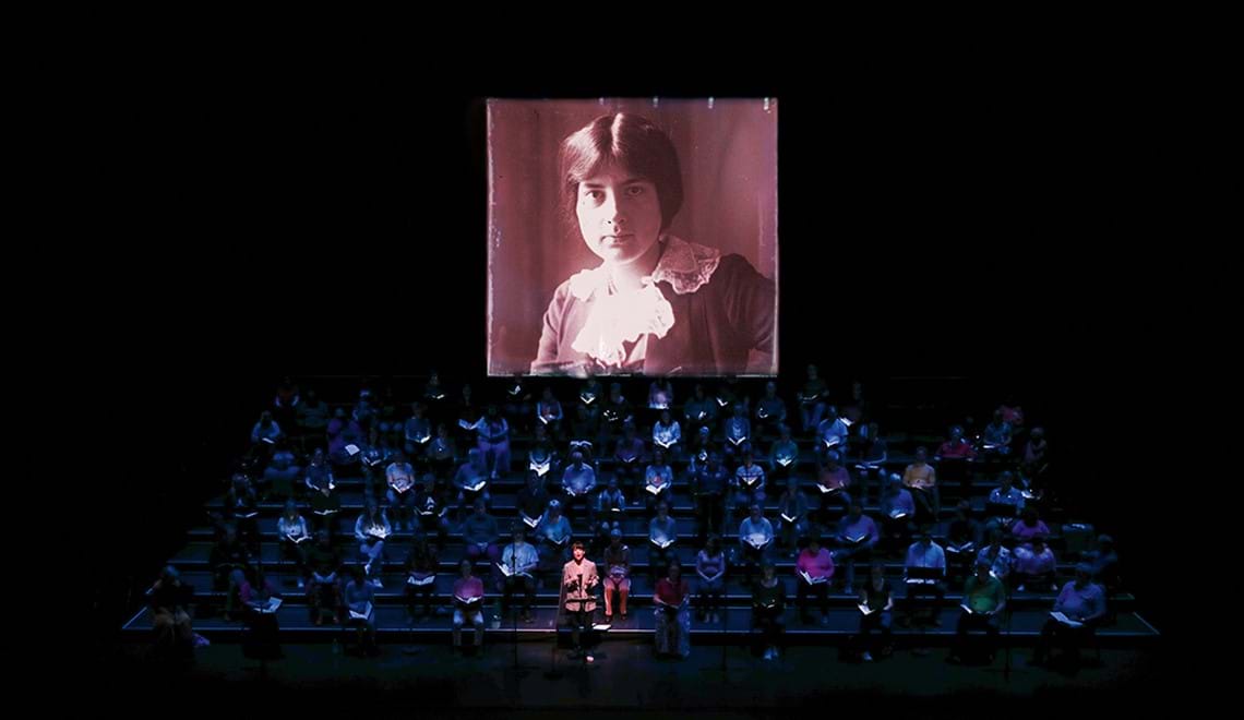 Singers and an orchestra sit on a dimly lit stage. An image of Lili Boulanger is projected behind them.