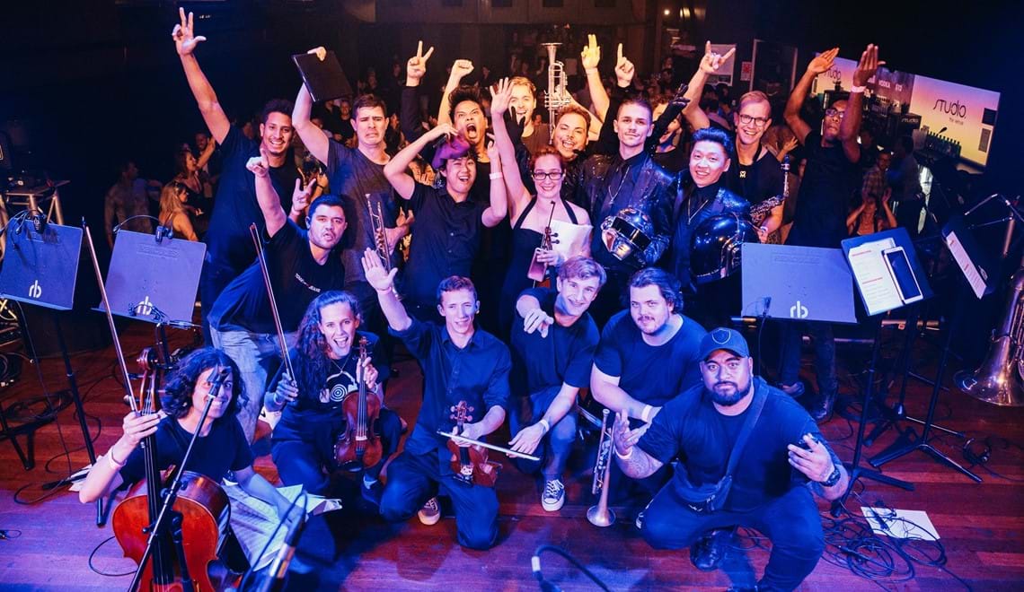The musicians of Alternative Symphony pose for the camera with their instruments. They are on a stage and an audience are cheering behind them.