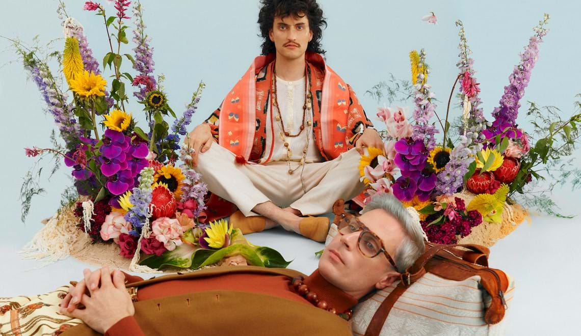 A photo of two men surrounded by flowers and wearing 70s-inspired outfits.