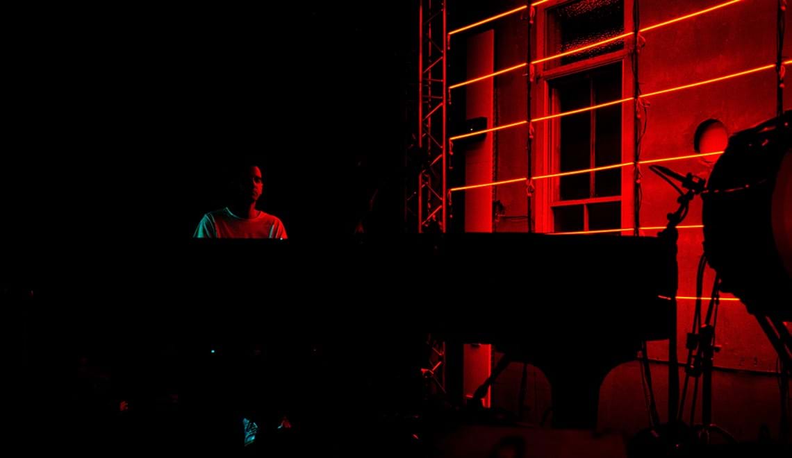 Josh Cohen sits at a piano in a darkened room. The lighting is red and dim, meaning Josh's face is only just visible.