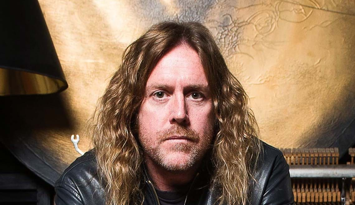 KRAM is seated and staring directly into camera with a serious expression. He has long, wavy blond ahair and is wearing a black leather jacket with ripped blue jeans.