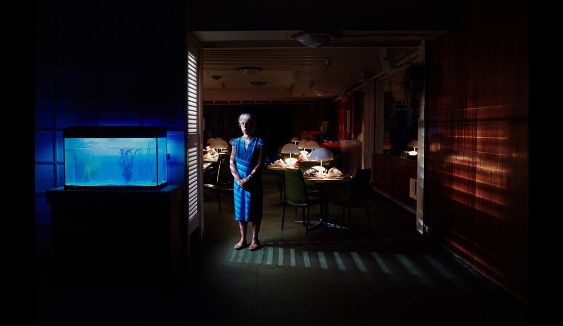 A woman in a blue dress stands in a dark room. To the left of her is a fish tank with blue lights and on the right is a room with rows of tables with rotary dial telephones on them