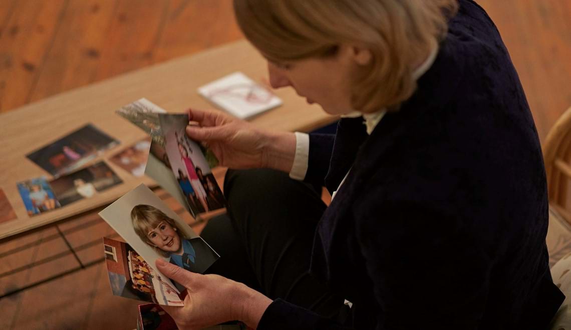 A woman sitting, looking at five pictures, one is a school photo, others of multiple children. A box is beside her and there are more pictures on the table in front of her