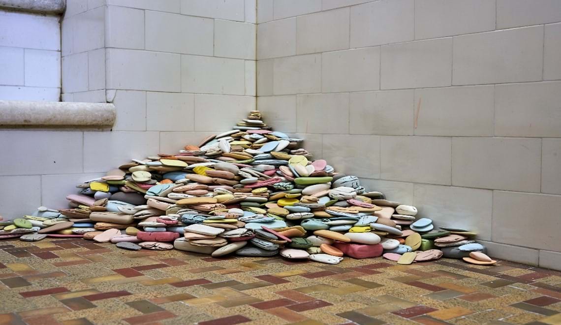 A pile of pastel coloured, worn down soap bars are pictured on a bathroom corner with brown floor tiles and white wall tiles 