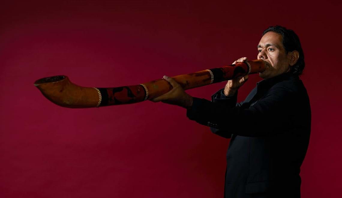 William Barton holds his didgeridoo to his mouth and into the air in front of a maroon background