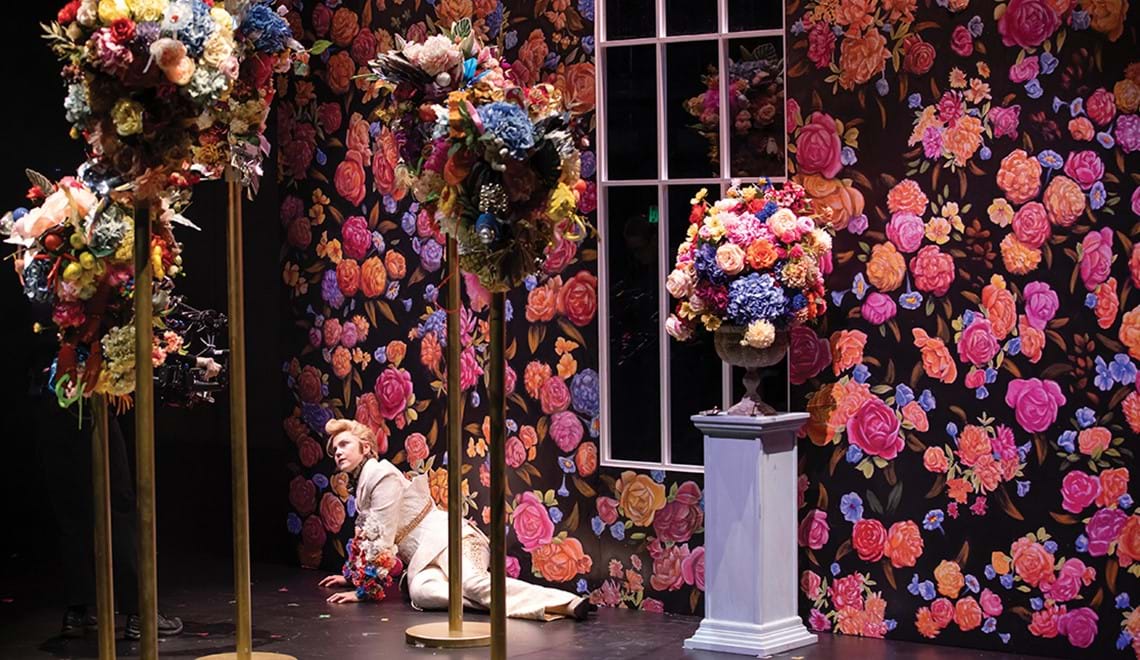 An actor sits on the ground in front of a wallpaper with large colourful flowers and surrounded by plinths topped by displays of flowers in similar colours.