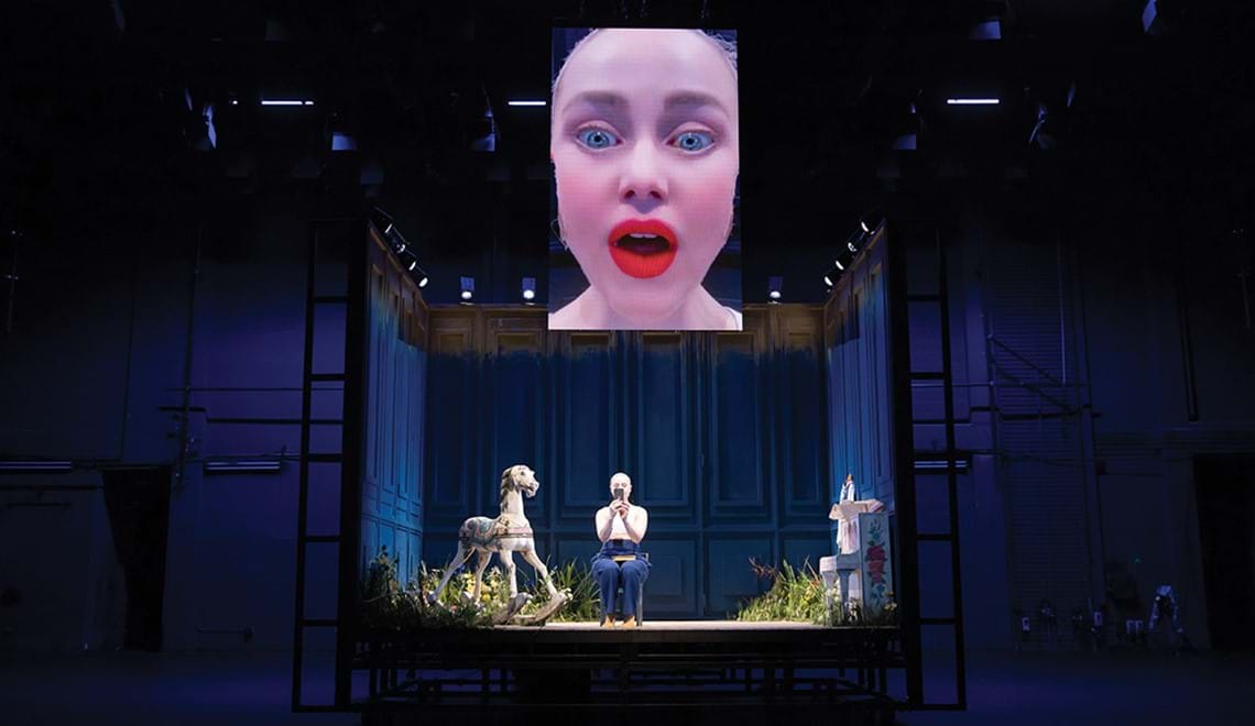 An actor sits in a box on a stage, holding a phone in front of their face. There is a screen above them showing their face (obviously coming from the phone's camera).