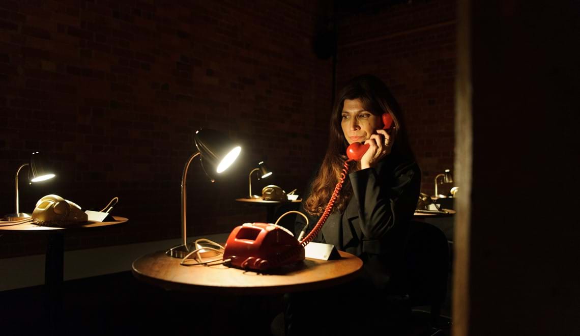 A woman in a black coat sits at a small round desk with a lamp and a phone on it. She is holding the receiver of a red rotary dial telephone to her ear.  Around her are more desks with rotary phones and lamps.