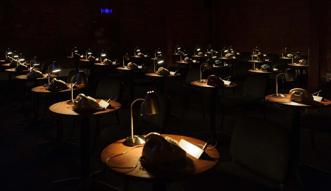 Rows of round desks are pictured in a dark room. On each a rotary dial telephone and a desk lamp.