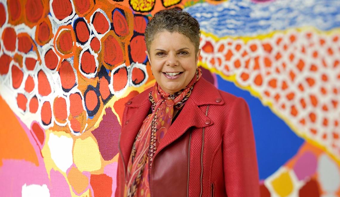 Deborah Cheetham wears a red jacket and red patterned scarf. She is smiling and standing in front of a painting by an Aboriginal artist.