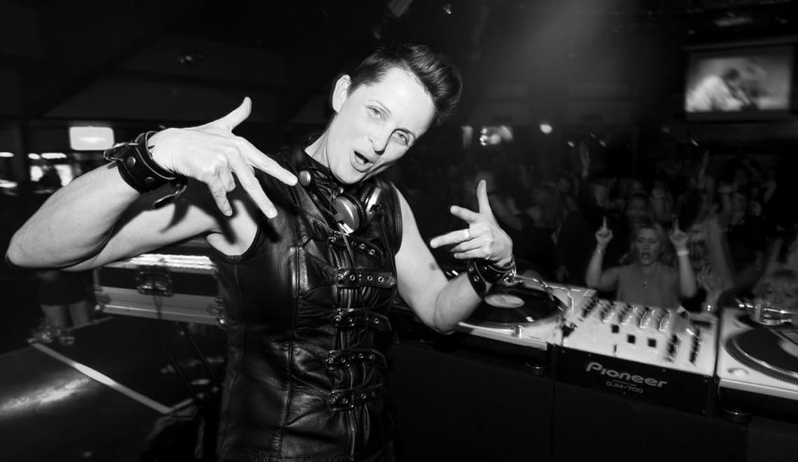 DJ JoSH is wearing a black leather vest and pulling a playfully tough expression while looking into the camera. Her DJ deck is behind her.