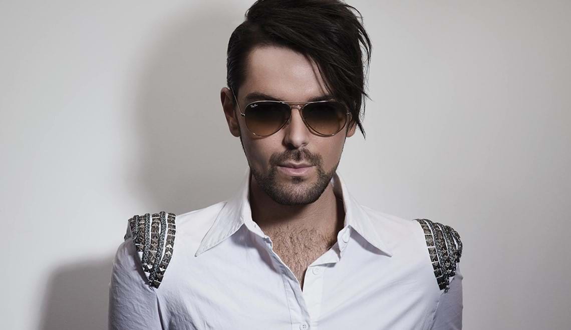 Filip wears a white shirt with embellished shoulders and sunglasses. He is facing the camera and looking directly into it.