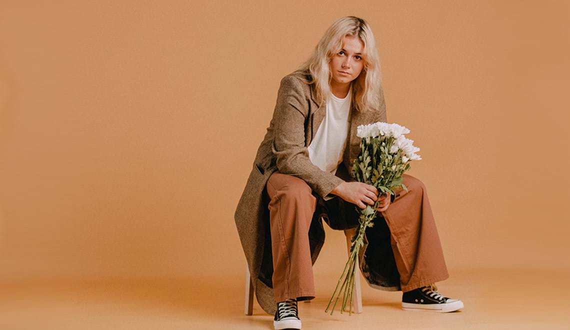Ruby Fields has blonde hair that falls to just below her shoulders. She is sittng on a chair and is dressed in a beige trenchcoat and rust-coloured pants with black Converse sneakers. She is holding a bunch of white flowers and is looking into the camera with a serious expression.