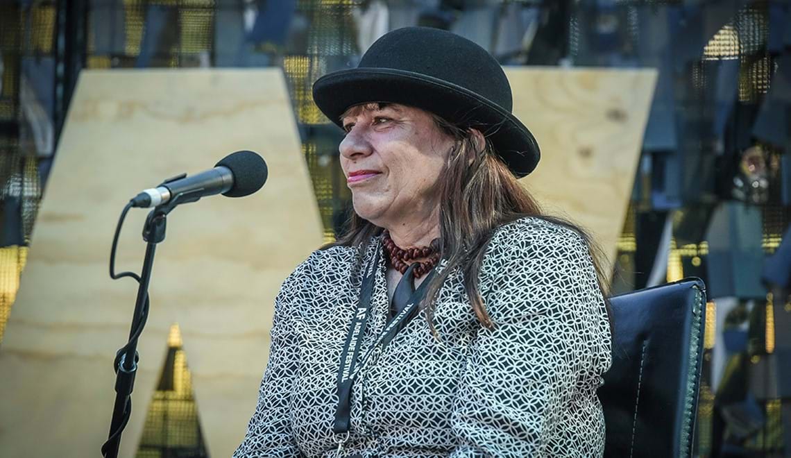 Ali Cobber Eckermann is pictured at 2020 Writers' Week. She is wearing a black and white shirt and a black hat. She is smiling slightly to the left of the camera and has a microphone in front of her.