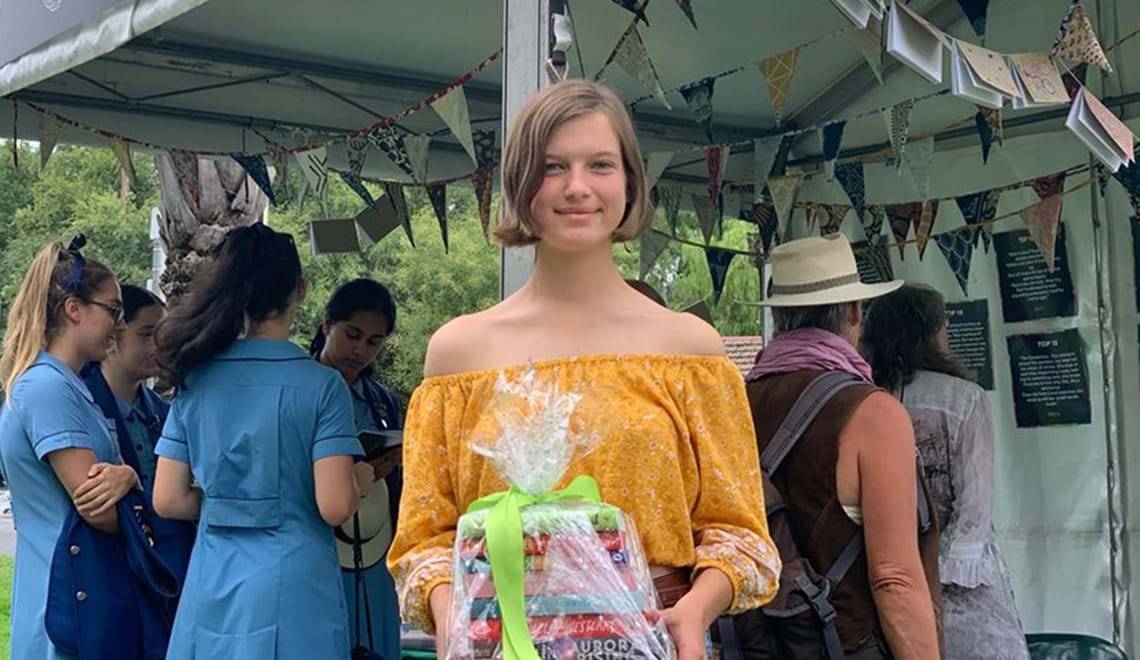 Emily Ashby, the 2020 winner of the microstory competition, has short, light brown hair and is wearing a yellow off-the-shoulder blouse. She is smiling at the camera and holding a stack of books wrapped in cellophane.