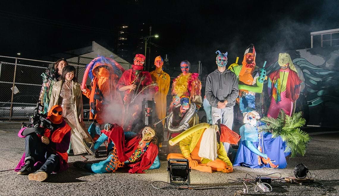 Members of The Bait Fridge wear brightly coloured costumes made of recycled materials. It's night time and they are posing outdoors in what appears to be an empty carpark.