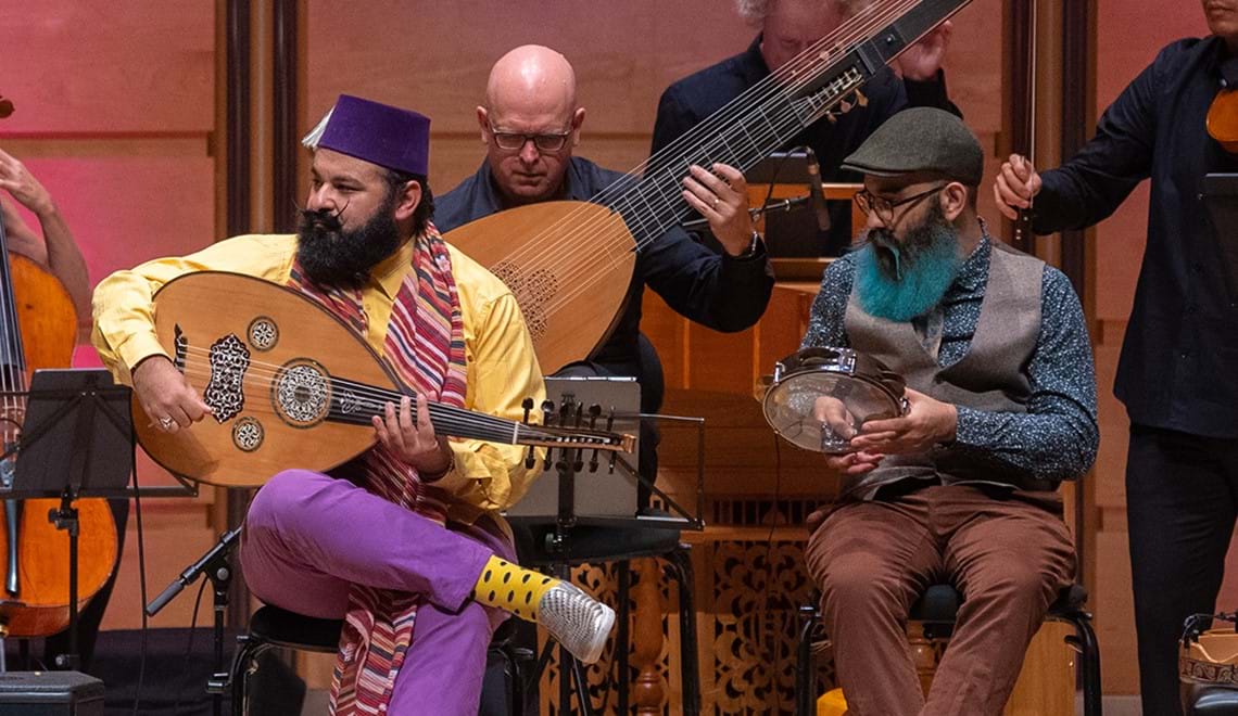 Joseph Tawadros is wearing a yellow shirt with purple pants, a purple hat and colourful scarf while holding an oud (string instrument). He is seated next to his brother, James Tawadros, who wears a peaked greay cap, a grey vest, brown pants and a teal shirt while holding a percussive instrument. Other musicians are visible behind them.