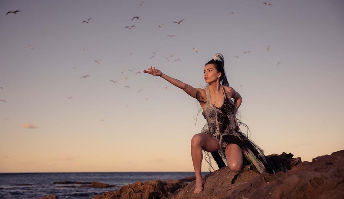 A woman crouches on a cliff face with one arm reaching out. The sea and a flock of flying birds are visible behind her.