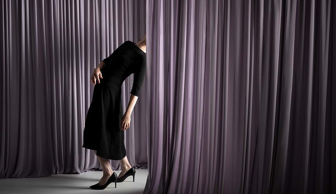 A woman wearing a black dress and heels is pictured leaning backwards. Her head is hidden behind a purple curtain.