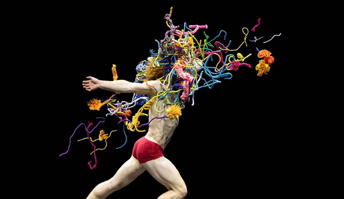 A man wearing red briefs poses in a lunge. His head is not visible as brightly coloured knitted objects are covering it - they look like they're exploding out from the spot where his head would be.