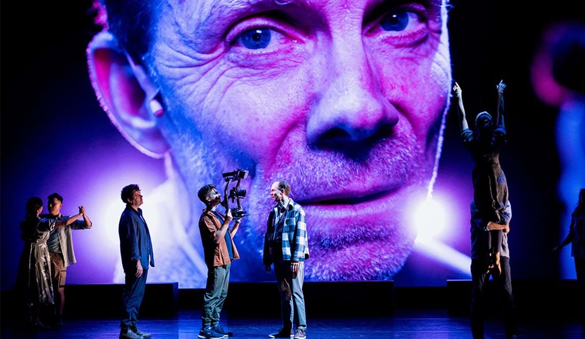 People in casual clothes stand on a stage in front of a screen with a close up of a mans face showing. One of the people on the stage are holding a large camera rig and other pairs are dancing together.