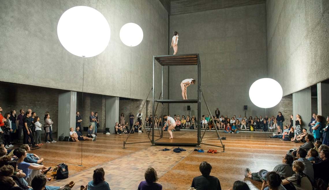 Three dancers are standing on a structure, similar to scaffolding, it has 3 levels and the dancers are wearing white t-shirts and no pants. There is a seated audience surrounding them.
