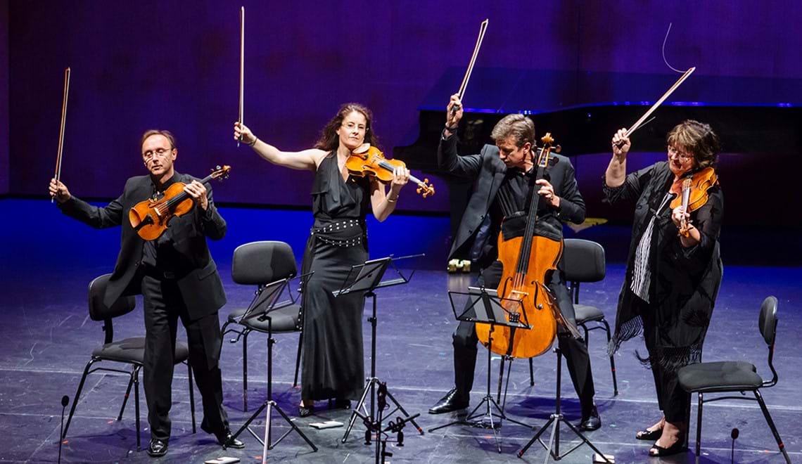 Four people, two men and two women, are wearing black and holding up their string instruments while standing on a stage.