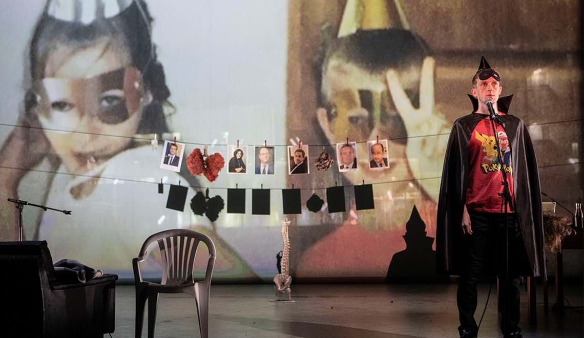 A man wearing a cape and a mask stands in front of a screen where photos of children dressed in a similar manner are projected. Photos are pegged onto strings in front of the screen.
