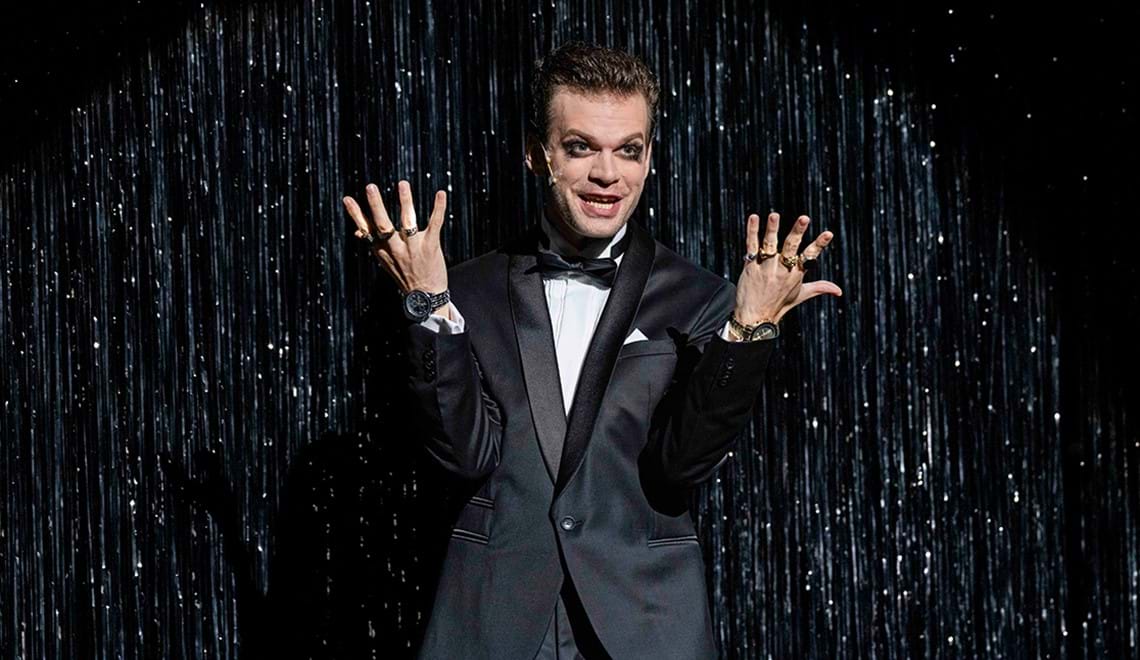 A man wearing a black suit and bow tie holds his hands up. He is wearing lots of large rings. The background is silver-spangled.