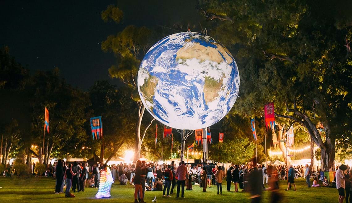 A lit up globe hovers above people walking on grass; trees and colourful flags are visible in the background.