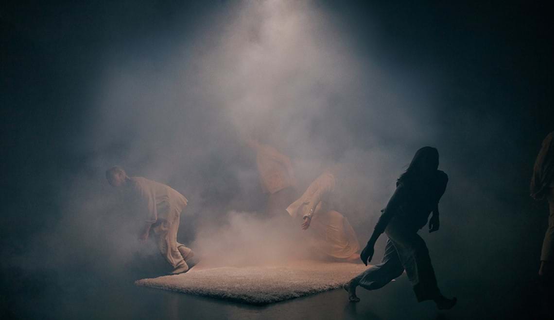Four people dressed in beige clothing are walking away from a shaggy white rug on a stage. There is a light with smoke coming over the rug, but the people are half in darkness.