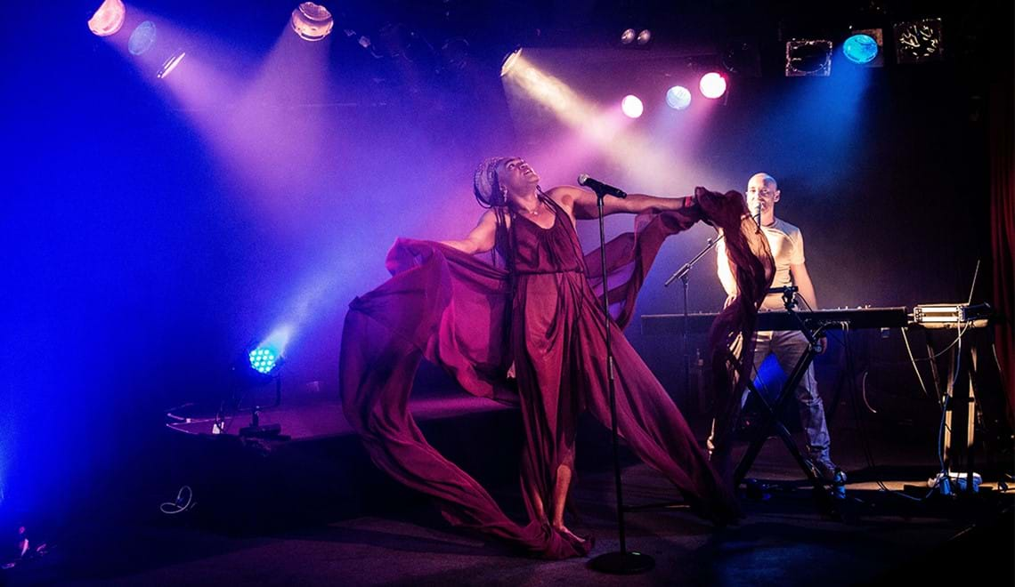 Two people stand on a stage in blue and pink light. One plays the keyboard and the other is wearing a flowing burgundy garment  with their arms held in the air.